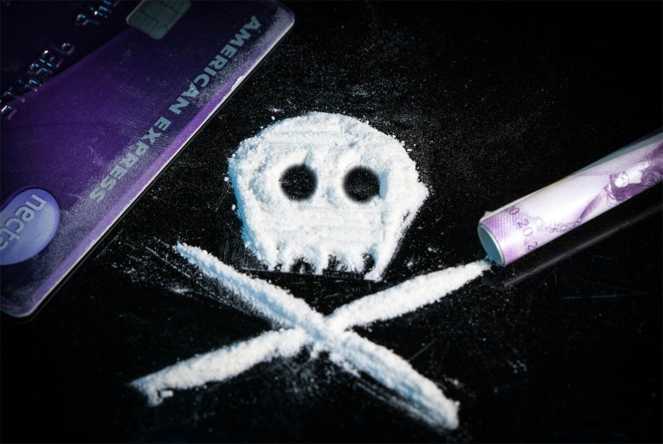 White powder arranged as a skull and crossbones beside a credit card and rolled banknote on a dark surface.