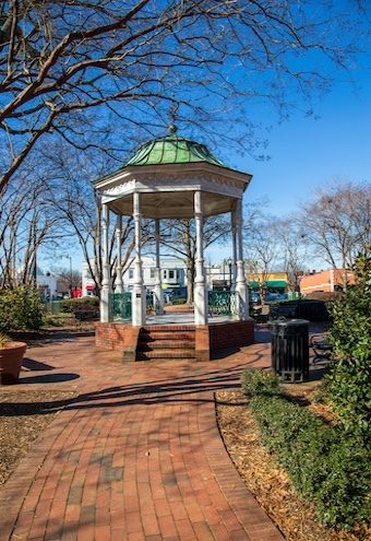 A white and green wooden gazebo on a red brick footpath surrounded by bare winter trees and lush green plants with a gorgeous clear blue sky at the Marietta Square in Marietta Georgia USA